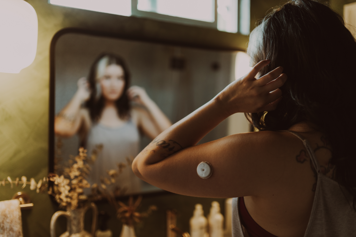 A person adjusts their hair while looking in a bathroom mirror. A continuous glucose monitor (CGM) is visible on their upper arm